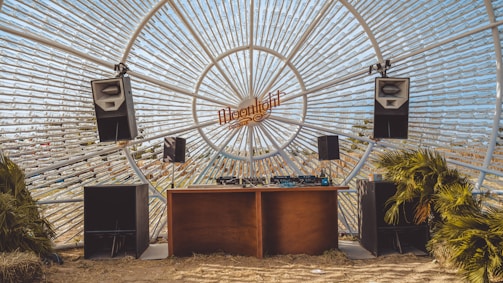 A unique DJ setup with large speakers and a wooden table under a dome of glass bottles. The backdrop creates a sunburst effect pointing outward, and there are some small palm plants placed nearby.
