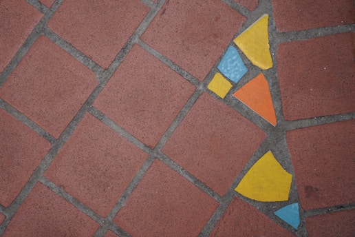 A brick-tiled surface with a cluster of colorful mosaic tiles embedded within. The mosaic features blue, yellow, and orange pieces arranged in a triangular pattern, providing a vivid contrast against the brownish-red bricks.