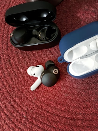 A pair of wireless earbuds in black are placed next to a similar pair in white on a textured red surface. Two open cases, one black and one blue, are visible nearby.