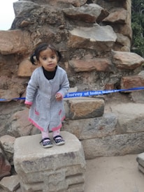 A young child with pigtails is standing on a stone platform, wearing a gray coat with pink accents, pink leggings, and black shoes. The setting includes a stone wall and a blue barrier tape in the background with 'Survey of India Delhi' partially visible.