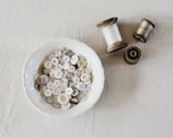 Rows of fine threads and buttons neatly arranged on an off-white wooden table.