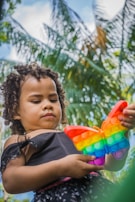 A child with curly hair is holding a colorful butterfly-shaped pop-it toy. Palm trees and blue sky create a natural, tropical backdrop. The child appears focused on the toy.