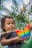 A child with curly hair is holding a colorful butterfly-shaped pop-it toy. Palm trees and blue sky create a natural, tropical backdrop. The child appears focused on the toy.