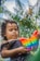 A child with curly hair is holding a colorful butterfly-shaped pop-it toy. Palm trees and blue sky create a natural, tropical backdrop. The child appears focused on the toy.