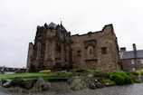 A historic stone building with arched windows and intricate masonry sits on a neatly tended lawn. The structure displays a medieval architectural style with strong, dark stone walls. In the foreground, there are rocks and various plants, adding a natural element to the scene. The sky is overcast, enhancing the classic ambiance of the location.