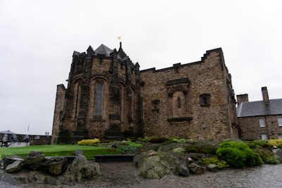 A historic stone building with arched windows and intricate masonry sits on a neatly tended lawn. The structure displays a medieval architectural style with strong, dark stone walls. In the foreground, there are rocks and various plants, adding a natural element to the scene. The sky is overcast, enhancing the classic ambiance of the location.