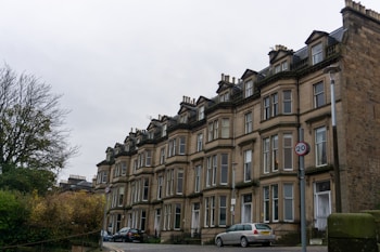 A row of classic British townhouses with bay windows and chimneys, set along a slightly curved street. The buildings are made of tan stone and feature multiple stories with a uniform architectural style. A couple of cars are parked on the street, and a 20 mph speed limit sign is visible. The sky is overcast, and a barren tree is to the left.