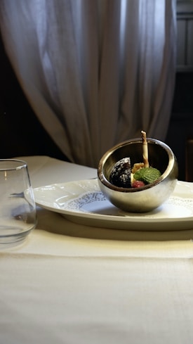 A metallic bowl with a dessert presentation is placed on a white plate with intricate patterns. The dessert includes fresh mint leaves, a small raspberry, and a breadstick. A glass is set on the table next to the plate, and soft drapes serve as the backdrop, creating a cozy ambiance.