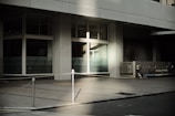 A modern building facade featuring large glass windows and concrete walls is captured in soft, natural lighting. The foreground shows a pavement with a metal post and chain barrier, and there is an air conditioning unit on the right side of the image.