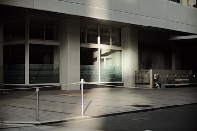 A modern building facade featuring large glass windows and concrete walls is captured in soft, natural lighting. The foreground shows a pavement with a metal post and chain barrier, and there is an air conditioning unit on the right side of the image.