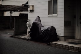 A motorbike is covered with a dark protective cover, parked against the side of a building in an urban setting. The surrounding area appears to be quiet with no people present, and the building has a tiled step leading to a door.