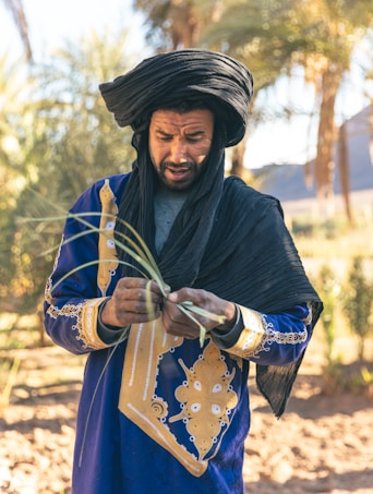 A man in traditional attire is focused on weaving or crafting something with green leaves. He wears a vibrant blue robe adorned with intricate gold patterns and a black head wrap. Palm trees and a blurred landscape are visible in the sunny background.