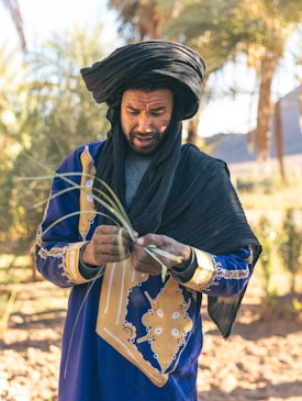 A man in traditional attire is focused on weaving or crafting something with green leaves. He wears a vibrant blue robe adorned with intricate gold patterns and a black head wrap. Palm trees and a blurred landscape are visible in the sunny background.