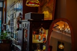 A cozy interior featuring a traditional wooden cabinet with ornate details, displaying bottles and other items on its shelves. A decorative mirror reflects a hanging chandelier and part of a warmly lit room. A plant in the background adds a touch of greenery, while various frames and decorations are arranged on the walls, including a whimsical holiday-themed ornament.
