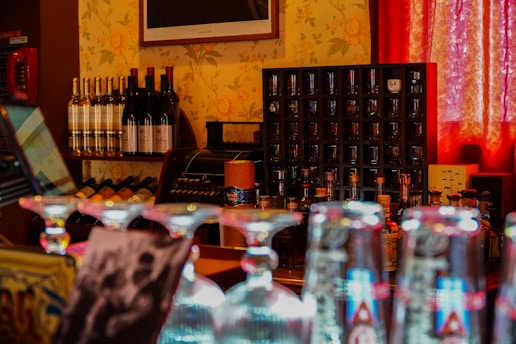 A cozy bar interior featuring a collection of wine bottles on a shelf, a lineup of liquor bottles, and several glassware pieces. A vintage cash register is displayed in the background, and the scene is warmly lit by soft red curtains.