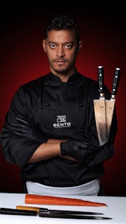 A serious-looking chef stands confidently with arms crossed, holding two large chef's knives. He is wearing a black chef's jacket with the logo 'Bento' and black gloves. In front of him on a white surface lies a large carrot and additional knives. The background features a bold, red gradient.