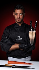 A serious-looking chef stands confidently with arms crossed, holding two large chef's knives. He is wearing a black chef's jacket with the logo 'Bento' and black gloves. In front of him on a white surface lies a large carrot and additional knives. The background features a bold, red gradient.
