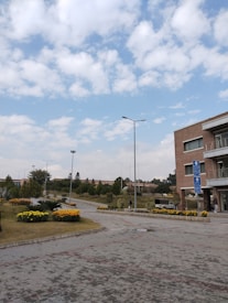 A well-maintained urban area with walkways, neatly trimmed bushes, and flower beds featuring yellow flowers. A modern brick building with balconies is on the right, and several street lamps line the pathways. The sky is partly cloudy with scattered patches of blue.