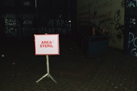 A small sign with the words 'AREA STERIL' in red letters is placed on a stand in a dimly lit area. The background features a wall with graffiti and various writings, creating a gritty urban atmosphere. There is a cart with two bare feet sticking out of it, suggesting a presence of a person. The setting is dark and conveys a sense of abandonment or neglect.