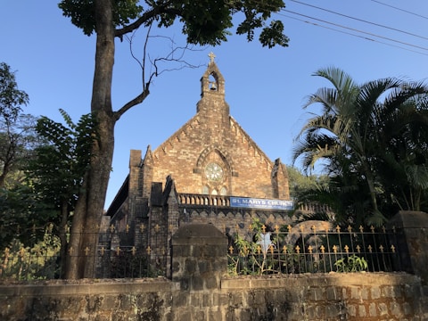 A stone church facade with a tall, narrow, triangular roofline features a large circular stained glass window. The building is surrounded by trees, including a tall tree to the left and a palm tree to the right. A wrought iron fence with decorative elements runs along the front. The sign above the entrance reads 'St. Mary's Church'. The lighting suggests a clear day with a blue sky.