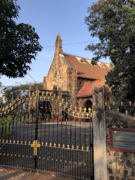A historic stone church with a red-tiled roof surrounded by lush green trees. An ornate black metal gate with gold accents featuring cross motifs stands in front. A sign on a pillar at the entrance provides information about the church.