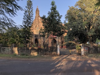 A rustic stone church is surrounded by lush trees and greenery. Its architecture includes arched windows and a prominent steeple. The church is enclosed by a stone wall and a gated entrance decorated with a golden cross.