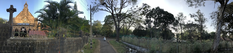 A quiet, narrow road leads past a church with a large cross at the entrance. The church is surrounded by greenery, including large palm trees and dense foliage. The scene is set against a backdrop of tall, mature trees, with a single person riding a bicycle down the path.