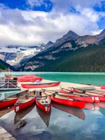 Several red canoes are lined up on a dock by a serene turquoise lake, surrounded by lush green forests and dramatic mountain peaks capped with snow under a partly cloudy blue sky.
