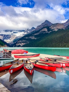 Several red canoes are lined up on a dock by a serene turquoise lake, surrounded by lush green forests and dramatic mountain peaks capped with snow under a partly cloudy blue sky.