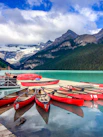 Several red canoes are lined up on a dock by a serene turquoise lake, surrounded by lush green forests and dramatic mountain peaks capped with snow under a partly cloudy blue sky.