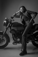 A woman in a black leather jacket and matching pants is leaning on a Royal Enfield motorcycle. The scene is captured in black and white, with the woman striking a confident pose. The lighting and shadows accentuate the textures of the leather and the machine.