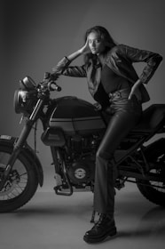 A woman in a black leather jacket and matching pants is leaning on a Royal Enfield motorcycle. The scene is captured in black and white, with the woman striking a confident pose. The lighting and shadows accentuate the textures of the leather and the machine.