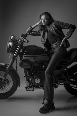 A woman in a black leather jacket and matching pants is leaning on a Royal Enfield motorcycle. The scene is captured in black and white, with the woman striking a confident pose. The lighting and shadows accentuate the textures of the leather and the machine.