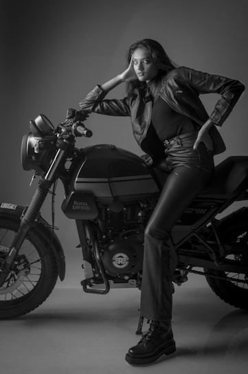 A woman in a black leather jacket and matching pants is leaning on a Royal Enfield motorcycle. The scene is captured in black and white, with the woman striking a confident pose. The lighting and shadows accentuate the textures of the leather and the machine.