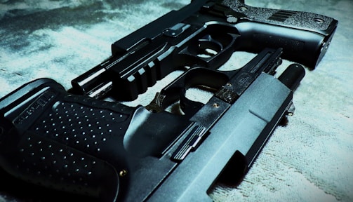 Two handguns are placed on a textured surface, one overlapping the other. The focus is on the intricate details of the gun surfaces, highlighting their metallic and textured grips.