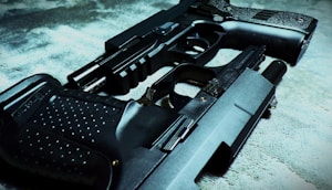 Two handguns are placed on a textured surface, one overlapping the other. The focus is on the intricate details of the gun surfaces, highlighting their metallic and textured grips.