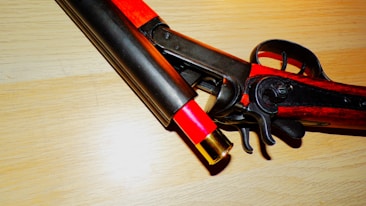 A close-up view of a break-action shotgun with a red shotgun shell partially inserted. The gun features dark metallic components and a wooden stock, placed on a light wooden surface.