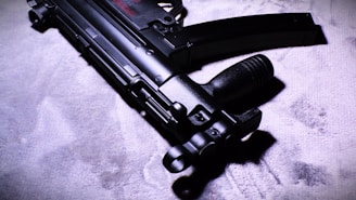 A close-up view of a sleek, black submachine gun placed on a textured surface. The image highlights the weapon's intricate details and matte finish under dramatic lighting, casting shadows and enhancing its features.