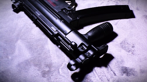 A close-up view of a sleek, black submachine gun placed on a textured surface. The image highlights the weapon's intricate details and matte finish under dramatic lighting, casting shadows and enhancing its features.