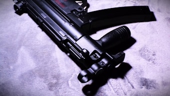 A close-up view of a sleek, black submachine gun placed on a textured surface. The image highlights the weapon's intricate details and matte finish under dramatic lighting, casting shadows and enhancing its features.