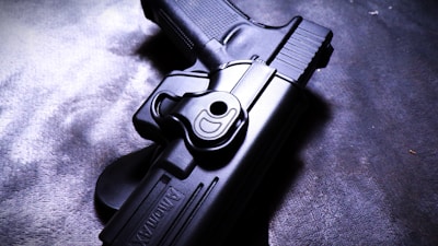 A close-up image of a handgun in a black holster lying on a textured surface. The firearm is partially visible, highlighting the details of the grip and trigger area. The lighting casts shadows, adding depth and contrast to the scene.