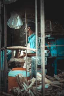 A man in a light blue shirt and apron stands in what appears to be a food or drink stall. The setting includes wooden structures, a large orange and blue cooler, and some kind of machinery or equipment. White napkins or cloths hang above, and the background seems to be an outdoor location with trees visible.