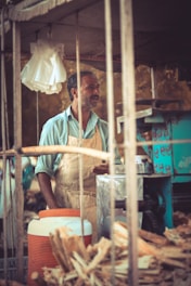 A person wearing a light blue shirt and apron stands behind a street vendor's cart, which contains a large thermos, sugarcane stalks, and a machine likely used for juice extraction. The setting appears rustic and outdoors, with a focus on manual labor and street food preparation.