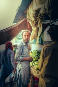 A man is standing in a market stall filled with fresh green vegetables, possibly cucumbers. He is wearing traditional clothing and seems to be engaging with someone or looking at something outside the frame. In the background, there are other people and market stalls visible under a canopy.