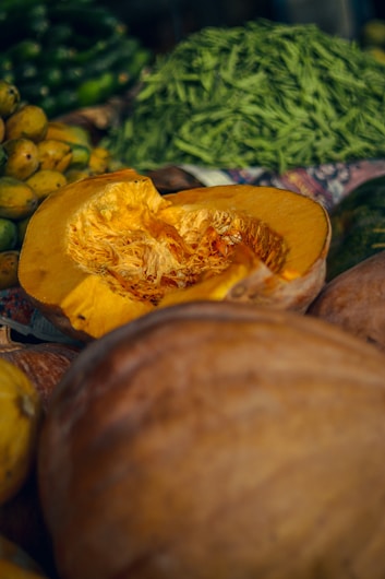 A close-up of a freshly cut pumpkin revealing its bright orange flesh and seeds. Surrounding the pumpkin are various green vegetables and fruits, including unripe mangoes, cucumber, and a pile of green beans in the background.