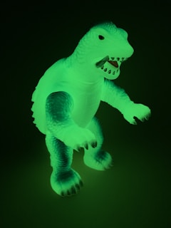 A glowing plastic dinosaur toy is visible in a dark setting. The figure has a textured body with embossed scales and an open mouth displaying sharp teeth. The dinosaur is standing upright with its arms slightly extended forward.