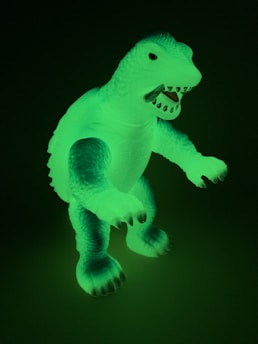 A glowing plastic dinosaur toy is visible in a dark setting. The figure has a textured body with embossed scales and an open mouth displaying sharp teeth. The dinosaur is standing upright with its arms slightly extended forward.