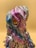 A sculpture or figurine with a surreal and abstract design, featuring exaggerated facial features and a wavy texture. The object displays a range of iridescent colors including pink, purple, and blue, giving it a metallic sheen. The eyes are distinct with a gear-like design and a yellow hue, contributing to a robotic or otherworldly appearance.