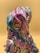 A sculpture or figurine with a surreal and abstract design, featuring exaggerated facial features and a wavy texture. The object displays a range of iridescent colors including pink, purple, and blue, giving it a metallic sheen. The eyes are distinct with a gear-like design and a yellow hue, contributing to a robotic or otherworldly appearance.