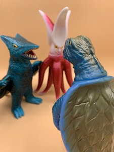 Three toy figures resembling monsters or creatures with unique features and colors are arranged on a beige background. One figure has a reptilian appearance with green scaly skin, another has a dragon-like appearance with blue and teal coloring, and the third features an octopus-like or alien character with a red body and white head.
